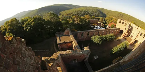 Eine alte Burgruine umgeben von grünen Hügeln. Im Innenhof sind einige Bäume und ein Parkplatz sichtbar.