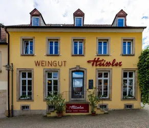 Ein charmantes Weingut mit gelber Fassade und großen Fenstern. Die elegante Beschilderung "Heißler" ist gut sichtbar und von Pflanzen umgeben.