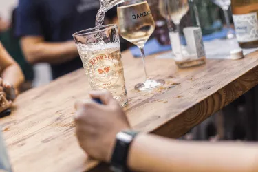 Ein Glas mit sprudelndem Getränk steht auf einer Holzbar. Im Hintergrund sind weitere Gläser und Menschen zu sehen.