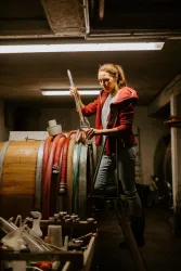 Eine Frau arbeitet in einem Weinkeller und steht auf einem Fass. Um sie herum sind verschiedene Werkzeuge und Schläuche zu sehen.