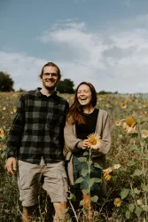 Ein Paar lächelt glücklich in einem Sonnenblumenfeld. Der Himmel ist blau mit ein paar Wolken.