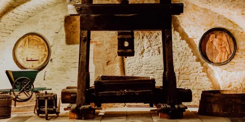 Ein historischer Weinkeller mit einer großen Holzpresse und Fässern. Die Wände sind mit kunstvollen Reliefs geschmückt.