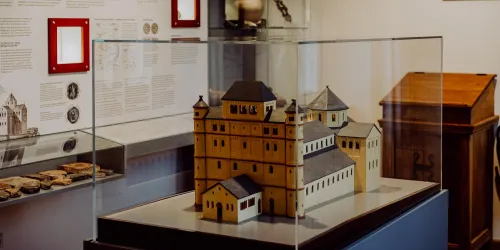 Ein detailliertes Modell eines historischen Gebäudes in einem Glaskasten. Im Hintergrund sind informative Tafeln und eine Holzkiste sichtbar.