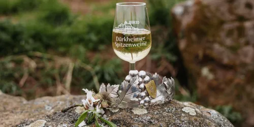 Ein Weinglas mit dem Aufdruck „Dürkheimer Spitzenweine“ steht auf einem Stein. Darunter befinden sich eine Silberkrone und frische Blumen.