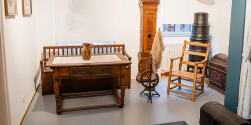 Ein historisch eingerichteter Raum mit einem Holzschreibtisch, einem Stuhl und einer Dekoration aus alten Möbeln. Im Hintergrund sind ein geschmiedeter Ofen und eine Wand mit Fenstern zu sehen.