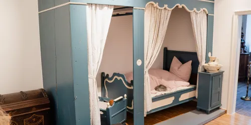 Ein stilvolles, blaues Kinderbett mit Vorhängen und Kissen. Neben dem Bett steht ein kleiner Nachttisch und im Hintergrund ist ein weiterer Raum sichtbar.