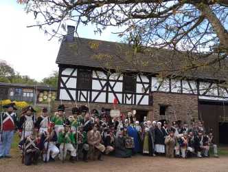 Eine Gruppe von Menschen in historischen Kostümen steht vor einem traditionellen Fachwerkhaus. Es scheint eine Nachbildung aus einer früheren Zeit zu sein, möglicherweise für eine Veranstaltung oder eine Reenactment-Darstellung.