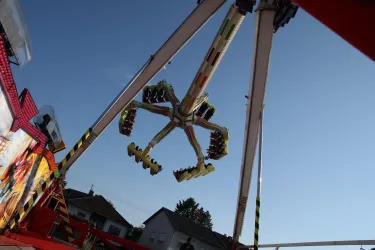Ein aufregendes Fahrgeschäft mit mehreren Armen, das sich gegen einen klaren blauen Himmel dreht. Im Hintergrund sind einige Häuser sichtbar.