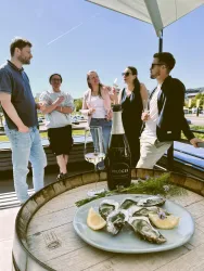 Eine Gruppe von fünf Personen genießt einen geselligen Moment im Freien. Auf einem Tisch liegt ein Teller mit Austern und eine Flasche Sekt.