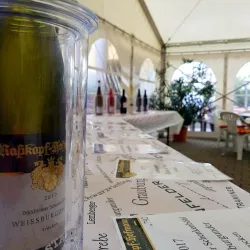 Ein Weinglas mit einer Flasche Weißburgunder in einem Kühlbehälter steht auf einem Tisch. Im Hintergrund sind mehrere Weingläser und eine dekorierte Tischdecke zu sehen.