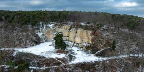 Eine schneebedeckte Felslandschaft mit einer felsigen Wand und kahlen Bäumen. Der Himmel ist bewölkt und verleiht der Szene eine ruhige Atmosphäre.