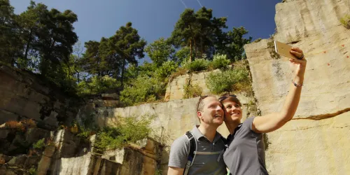 Ein Paar macht ein Selfie in einer malerischen Steinbruchlandschaft. Im Hintergrund sind hohe Felsen und Bäume zu sehen.
