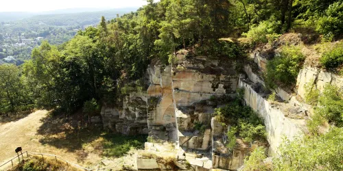 Eine beeindruckende Felslandschaft umgeben von Bäumen. Im Hintergrund ist eine Stadt sichtbar, die durch die Natur leicht versteckt wird.
