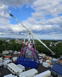 Ein großer Fahrgeschäft mit einem weißen und pinken Gestell steht auf einem Veranstaltungsgelände. Im Hintergrund sind Wolken und ein Teil der Stadt sichtbar.