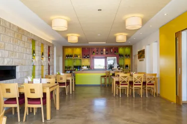 Ein modernes Restaurant mit Holztischen und bunten Wänden. Die Bar im Hintergrund ist mit Flaschen dekoriert und sorgt für ein einladendes Ambiente.