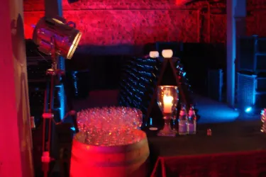 Eine stilvolle Eventlocation mit schummriger Beleuchtung. Im Vordergrund stehen Gläser auf einem Holzfass und eine ansprechend dekorierte Bar.