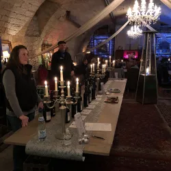 Ein festlich gedeckter Tisch mit Weinflaschen und Kerzen in einem stilvollen Raum. Im Hintergrund stehen Menschen und eine elegante Beleuchtung schafft eine gemütliche Atmosphäre.