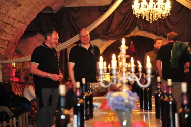 Eine gemütliche Weinstube mit einem langen Tisch, der mit Weinflaschen und Kerzen dekoriert ist. Im Hintergrund sind mehrere Personen, die sich unterhalten.