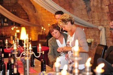 Zwei Frauen betrachten Weingläser in einem romantischen, beleuchteten Weinkeller. Im Hintergrund sind Kerzen und Weinfässer sichtbar.