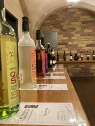 Eine Reihe von Weinen steht auf einem Holzregal. Im Hintergrund sind Steine und Flaschen zu sehen, die eine gemütliche Atmosphäre schaffen.