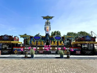Ein farbenfroher Freizeitparkstand mit aufregenden Fahrgeschäften und dekorativen Elementen. Im Hintergrund blauer Himmel und grüne Bäume.