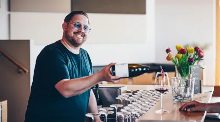 Ein lächelnder Mann steht an einer Theke und hält eine Flasche Wein. Im Hintergrund sind Gläser und eine Vase mit Tulpen sichtbar.