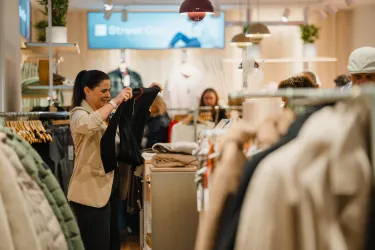 Eine Frau hält ein Kleidungsstück in einem modernen Bekleidungsgeschäft. Im Hintergrund sind weitere Kunden und eine ansprechende Verkaufsumgebung zu sehen.