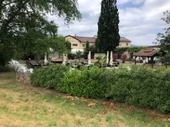 Ein gemütlicher Außenbereich mit Tischen und Stühlen, umgeben von Bäumen und Sträuchern. Im Hintergrund ist ein Gebäude sichtbar, das eine entspannte Atmosphäre ausstrahlt.