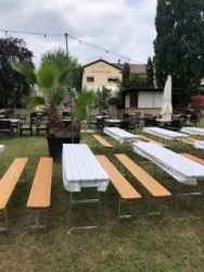 Ein Außenbereich mit langen Holztischen und Bänken. Es gibt einige Pflanzen und eine entspannte Atmosphäre.