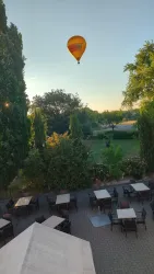 Ein Heißluftballon schwebt über einem grünen Garten. Darunter stehen Tische und Stühle in einem gemütlichen Außenbereich.