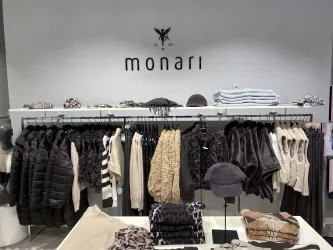 Ein modernes Geschäft mit Kleidung der Marke Monari. Auf den Regalen hängen verschiedene Oberbekleidungsstücke und Accessoires in neutralen Farben.