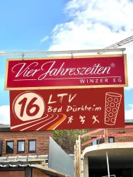 Ein großes Schild mit dem Schriftzug "Vier Jahreszeiten WINZER EG" und der Nummer "16 Haardtblick". Der Hintergrund zeigt einen klaren Himmel und ein neugebautes Gebäude.