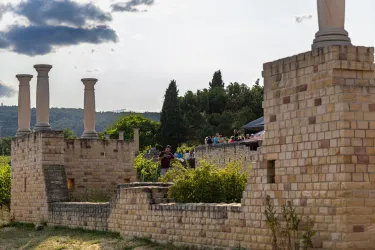 Eine historische Ruine mit Säulen und Mauern, umgeben von üppigem Grün. Im Hintergrund sind Menschen zu sehen, die die Aussicht genießen.