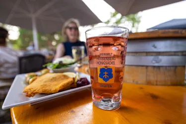 Ein Glas mit einer pinkfarbenen Getränk steht auf einem Holztisch. Im Hintergrund ist ein Teller mit Essen und eine Person zu sehen.