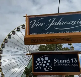 Ein auffälliges Schild der „Vier Jahreszeiten Winzer EG“ mit dem Hinweis „Tempel“. Im Hintergrund ist ein Riesenrad und eine rustikale Holzkonstruktion zu sehen.