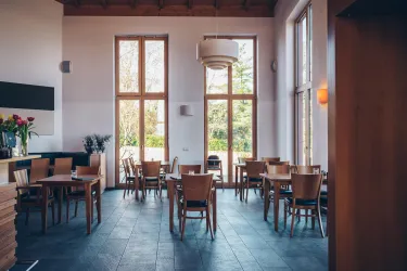 Ein modernes Restaurant mit Holztischen und Stühlen. Große Fenster lassen viel Licht herein und bieten einen Blick ins Grüne.