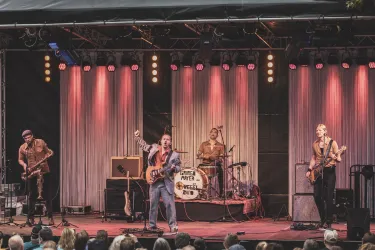 Eine Live-Band spielt auf einer Bühne mit bunten Lichtern. Die Musiker treten energisch auf, während das Publikum zuschaut.