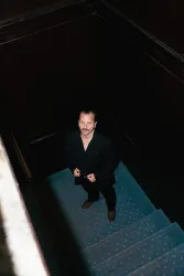 Ein Mann in einem schwarzen Anzug steht auf einer Treppe. Der Raum hat eine dunkle Atmosphäre mit blauen Teppichen.