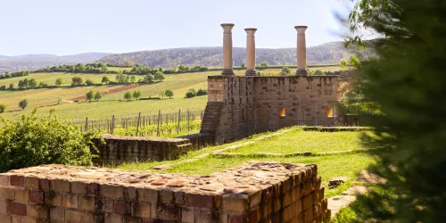 Eine alte Ruine mit mehreren Säulen steht in einer grünen Landschaft. Im Hintergrund sind sanfte Hügel und Weinreben zu sehen.