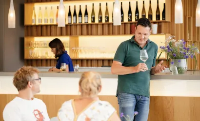 Ein Mann erklärt etwas an einer Bar, während zwei Gäste aufmerksam zuhören. Im Hintergrund ist eine Frau, die Getränke vorbereitet.