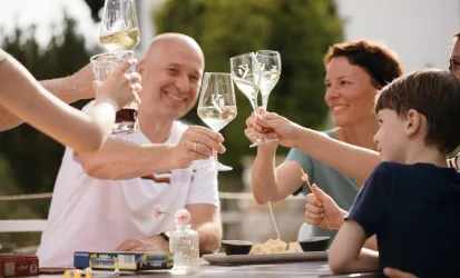 Eine Gruppe von Personen stößt mit Gläsern an und genießt eine gesellige Zeit im Freien. Auf dem Tisch sind Snacks und Getränke zu sehen.