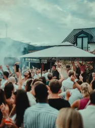 Eine lebhafte Party im Freien mit einer großen Menschenmenge. Der DJ spielt Musik unter einem Pavillon, während die Gäste feiern und ihre Hände heben.