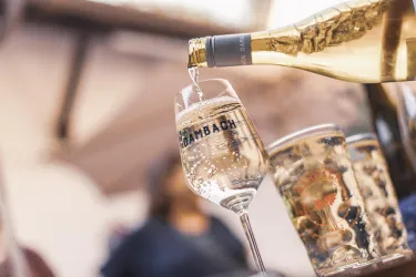 Ein Glas feiner Schaumwein wird gerade eingeschenkt. Im Hintergrund sind verschwommen weitere Gläser zu sehen.