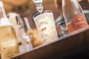 Ein Glas mit sprudelndem Getränk steht auf einem Tisch, umgeben von Flaschen. Der Fokus liegt auf dem Glas mit dem Dambach-Logo.