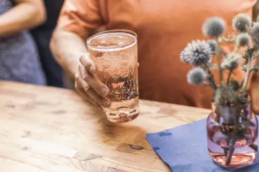 Ein Glas mit einem sprudelnden Getränk wird von einer Person gehalten. Auf dem Tisch steht auch eine kleine Vase mit Blumen.