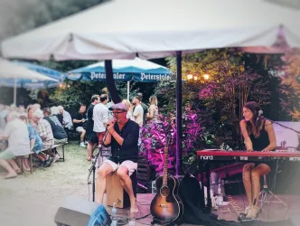 Ein Live-Musikauftritt mit einem Sänger und einer Pianistin unter einem Pavillon. Im Hintergrund sind eine Gruppe von Menschen und Tische zu sehen.