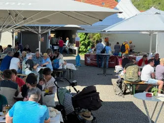 Ein belebter Außenbereich mit vielen Menschen, die an Tischen sitzen und essen. Im Hintergrund sind Stände mit Speisen und Getränken sichtbar.