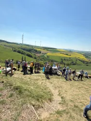 Eine Gruppe von Menschen versammelt sich auf einer Wiese mit grünen Hügeln und Windkraftanlagen im Hintergrund. Der Himmel ist klar und blau, was einen sonnigen Tag zeigt.
