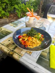 Ein Teller Pasta mit buntem Gemüse und frischen Kräutern. Daneben steht ein Glas Weißwein auf einem stilvollen Tisch.