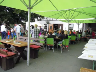 Ein gemütlicher Außenbereich mit vielen Gästen unter grünen Sonnenschirmen. Tische sind mit bunten Stühlen umgeben, was eine einladende Atmosphäre schafft.